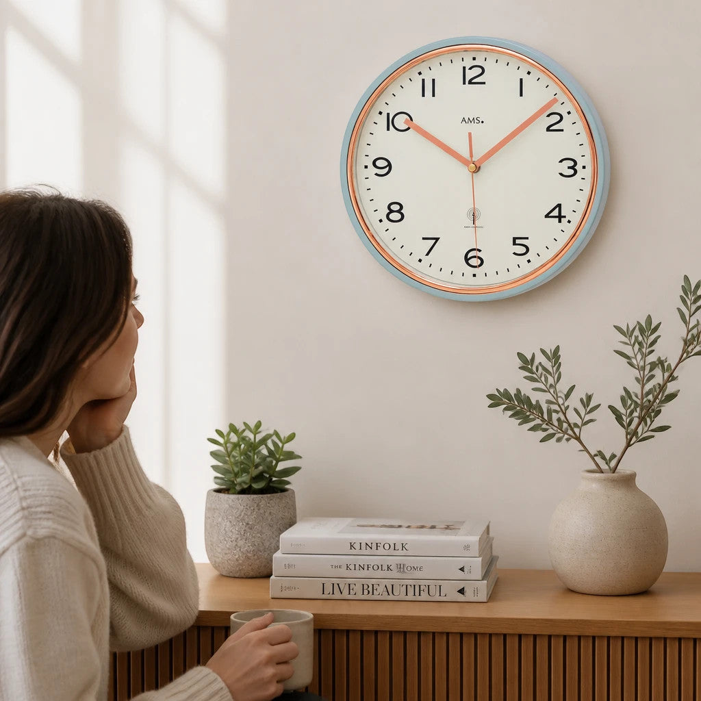 Standuhren.com 🕰️ AMS Funkwanduhr mit hellblauem Metallgehäuse, goldenem Zierring und weißem Zifferblatt an einer Wand.